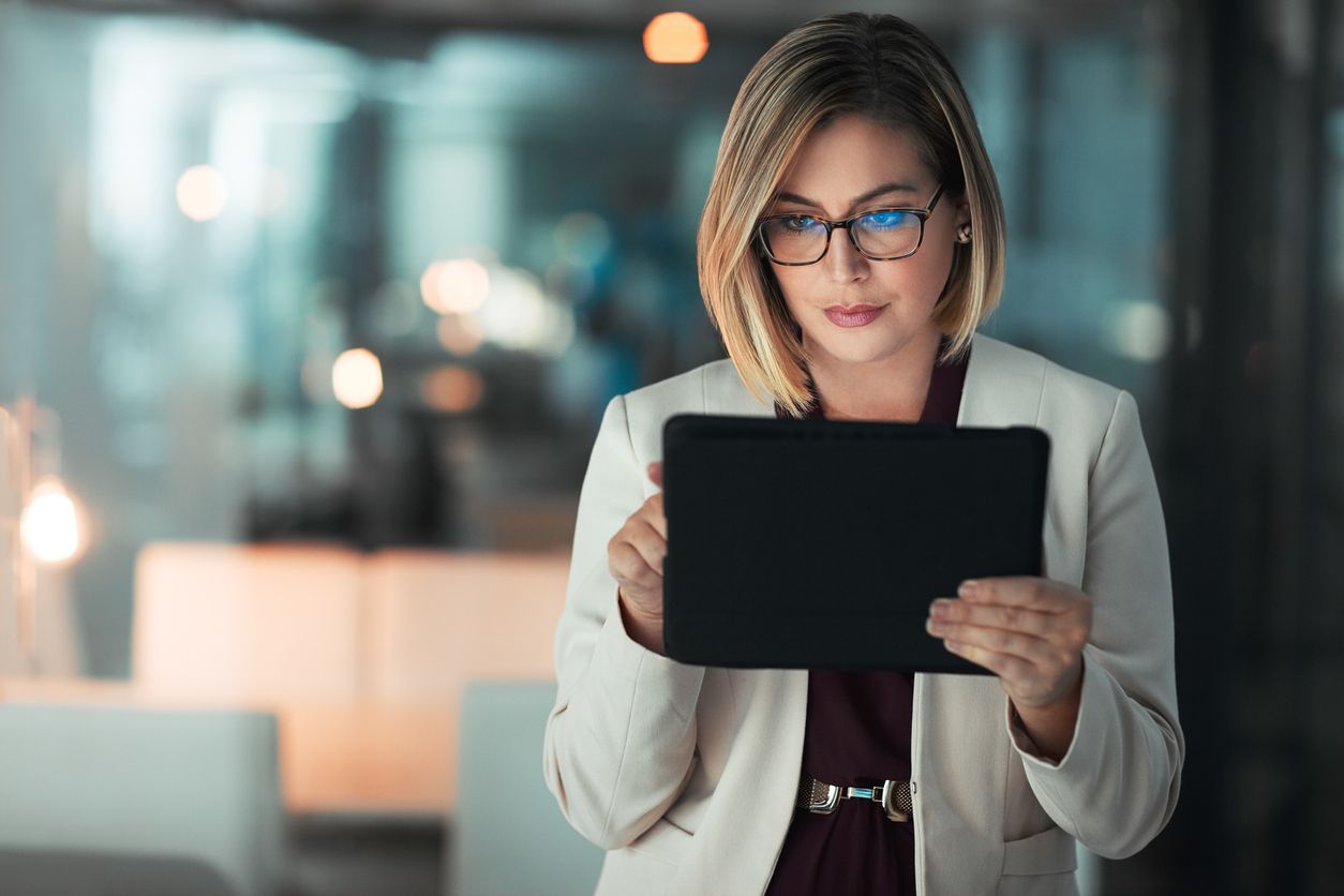A woman in glasses focused on a tablet, with blue light reflecting from its screen onto her lenses.