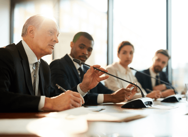 Four professionals sit at a conference table with individual microphones, one speaking.