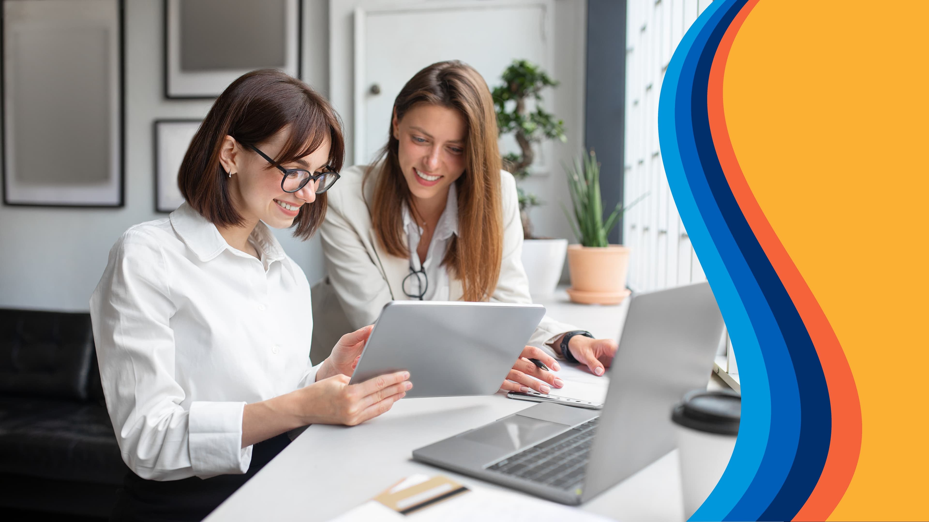 two women are sitting at a table looking at a tablet and a laptop .