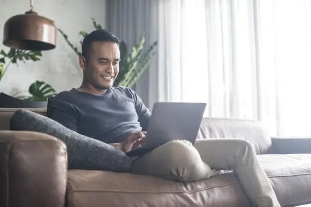 A smiling man sits on a brown sofa, using a laptop.