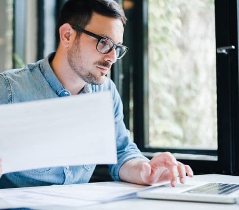 Man with glasses reviewing documents and typing on a laptop.