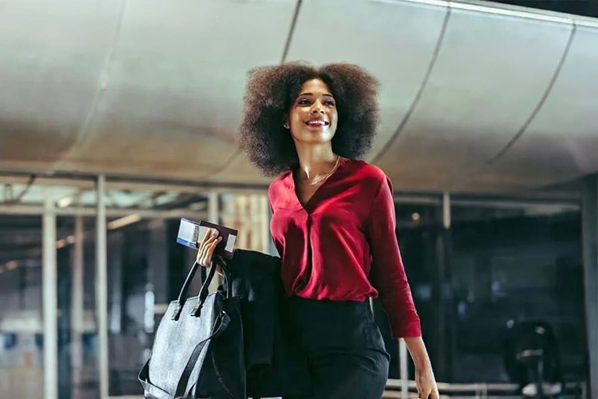 A smiling woman with an afro hairstyle, wearing a red blouse, carries a handbag and travel documents in an airport.