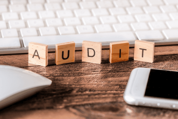 Wooden letter tiles spelling AUDIT on a desk with a keyboard, mouse, and smartphone.