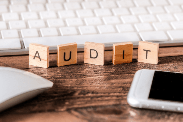 Wooden letter tiles spelling AUDIT on a desk with a keyboard, mouse, and smartphone.