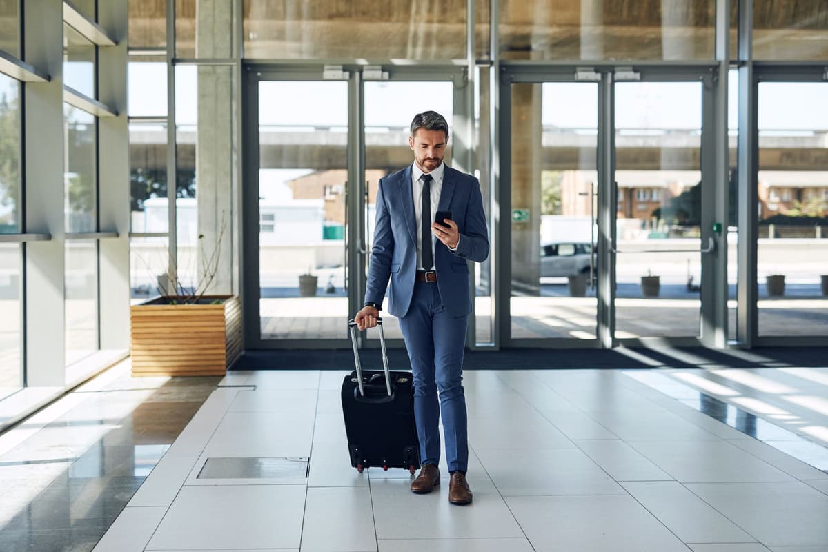 A man in a blue suit pulls a black suitcase while looking at his phone in a modern lobby.