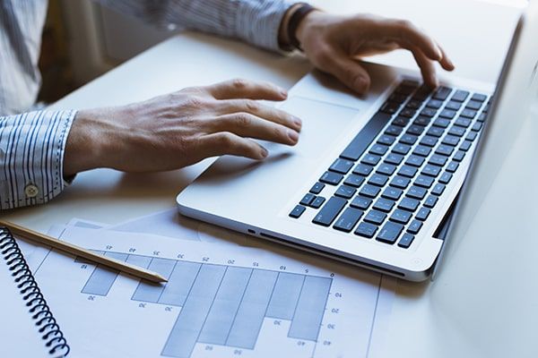Hands typing on a laptop, next to a bar graph and pencil on a desk.