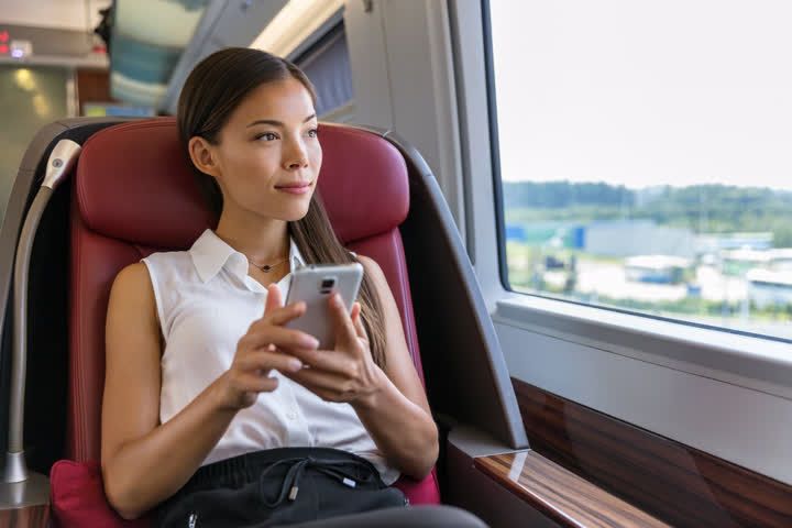 A woman in a train seat, holding a phone and looking out the window.