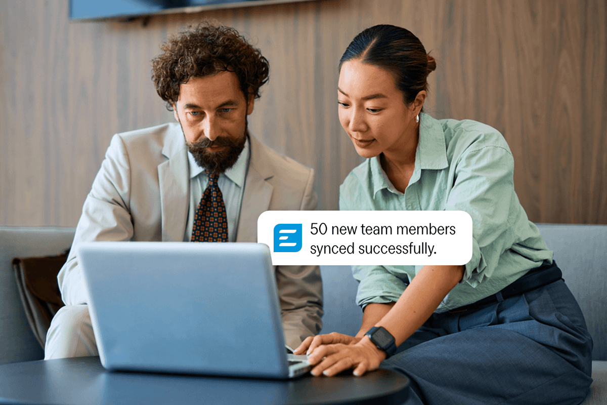 Two professionals look at a laptop displaying a notification that reads, "50 new team members synced successfully."