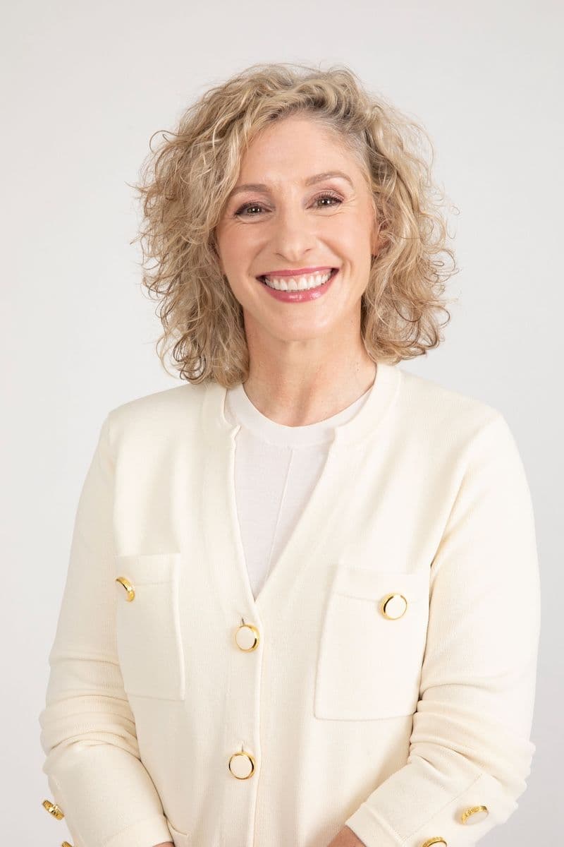 A smiling woman with curly blonde hair wears a cream cardigan with gold buttons.