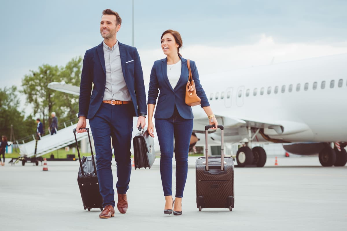 A smiling man and woman walk with luggage on an airport tarmac next to an airplane.