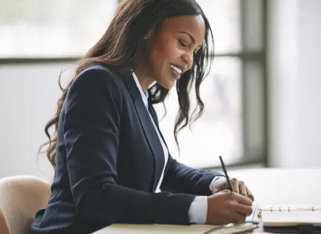 une femme en costume est assise à un bureau et écrit sur un morceau de papier.