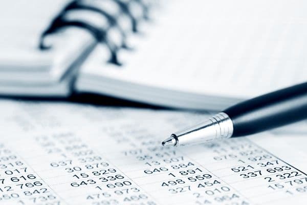 A pen rests on a document filled with numbers, with a spiral notebook in the blurred background.