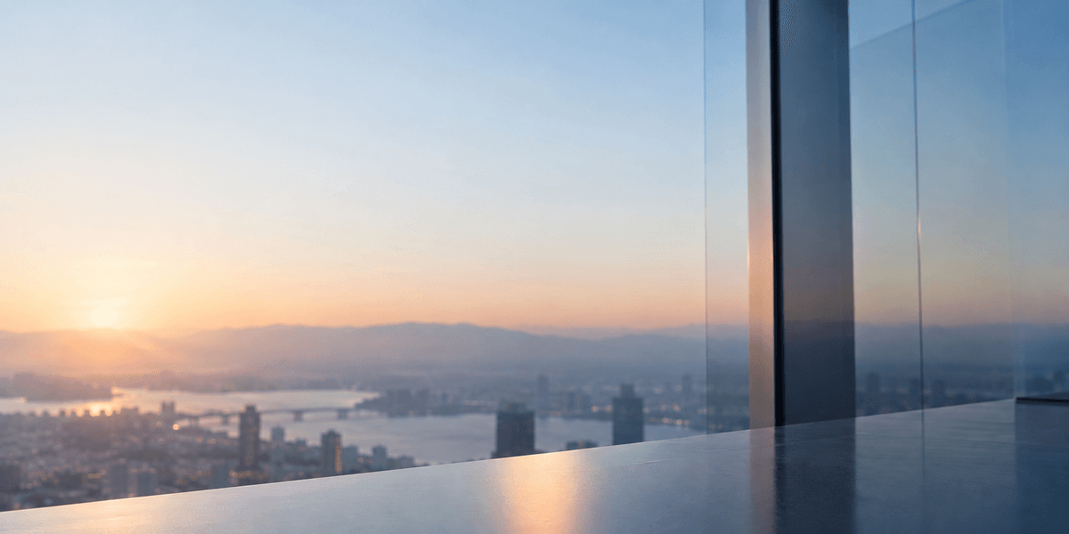Sunset cityscape with a river and distant mountains, viewed from a high-rise window with reflections.