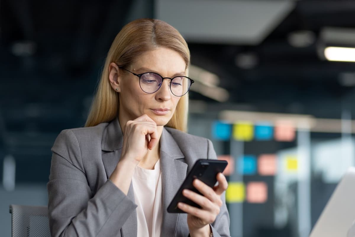 A thoughtful blonde woman in glasses and a suit looks at her phone.