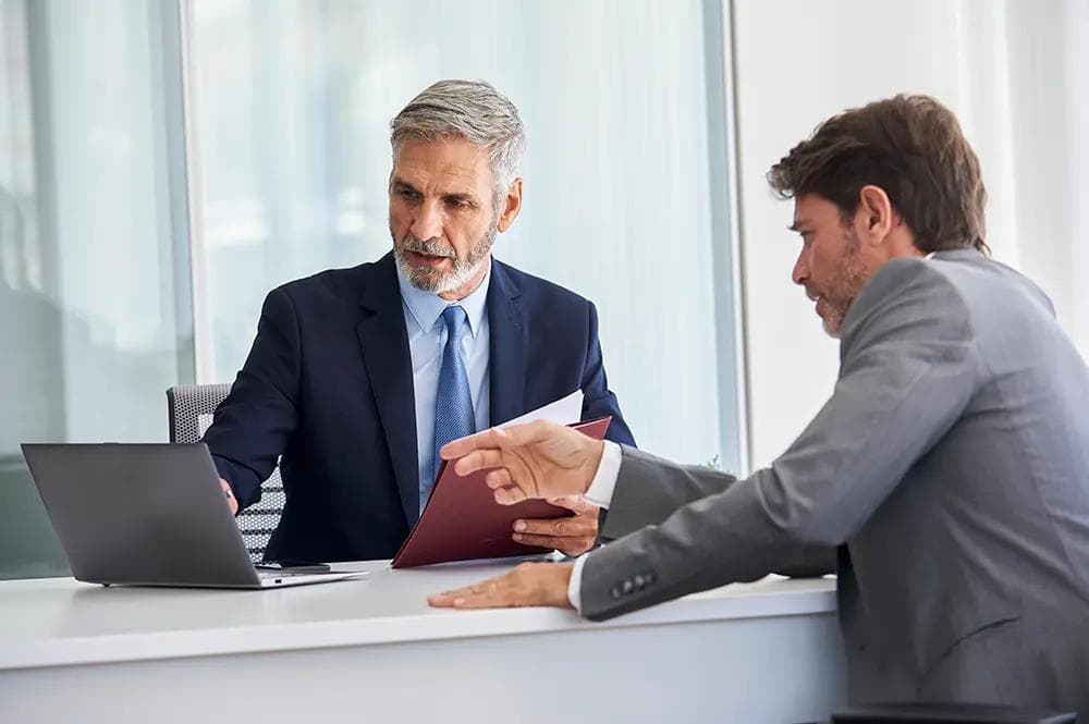 Two businessmen discuss documents and a laptop at a desk.