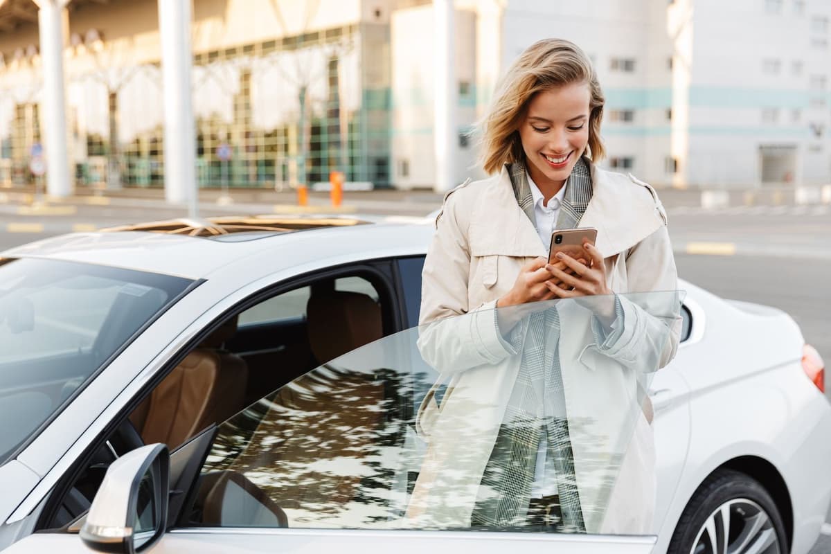 A smiling woman in a trench coat checks her phone while leaning on a white car.