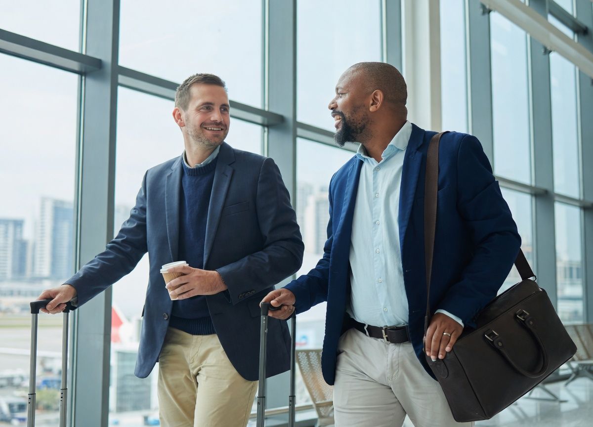 Two well-dressed men smiling and conversing while walking with luggage in an airport.