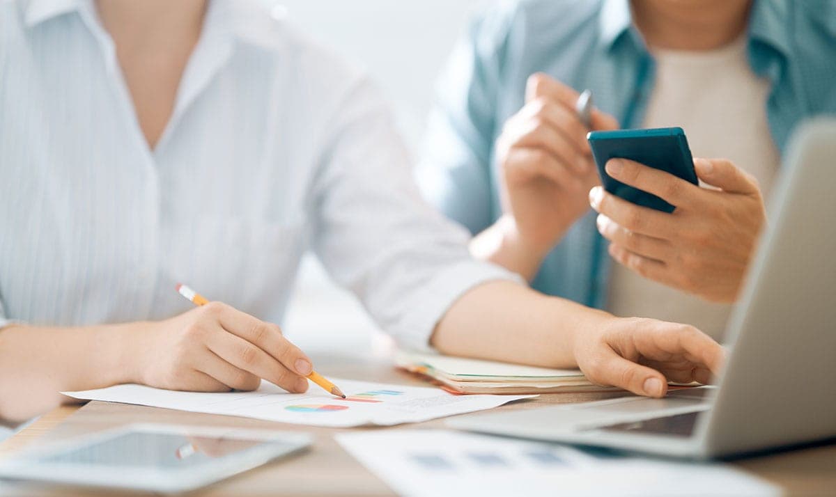 Two people collaborating at a desk with documents, a laptop, and a smartphone.