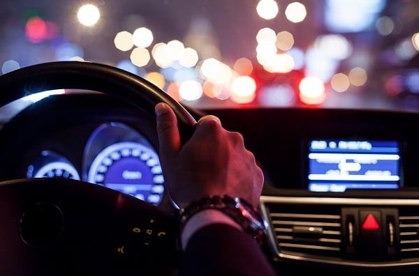 Hand on a steering wheel, driving at night with blurred city lights.