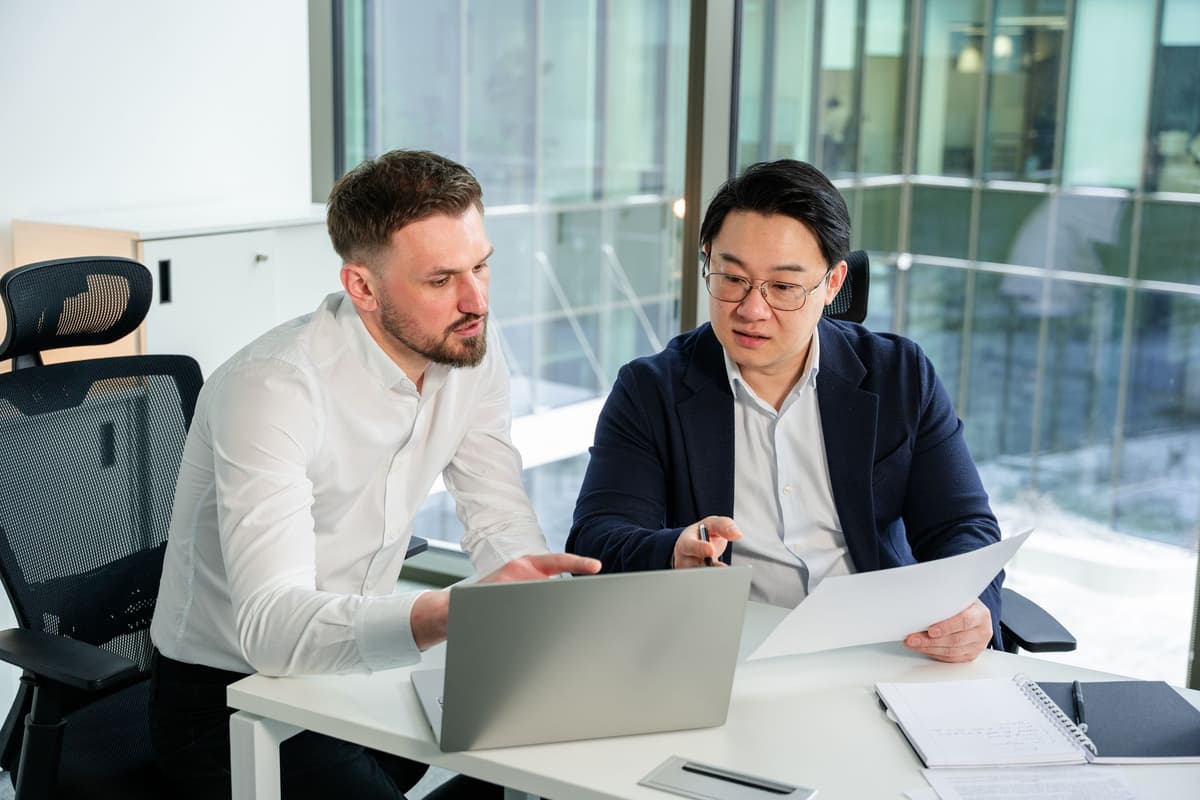 Two men in an office collaborating over a laptop, one holding papers and pointing.