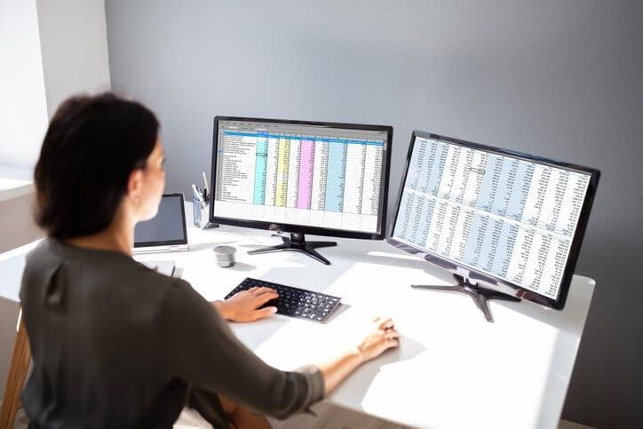 Woman working at a desk with two monitors showing spreadsheets, one in portrait mode.