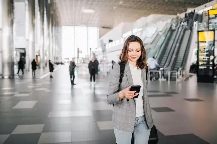 A young woman checks her phone while walking through an airport terminal with luggage.