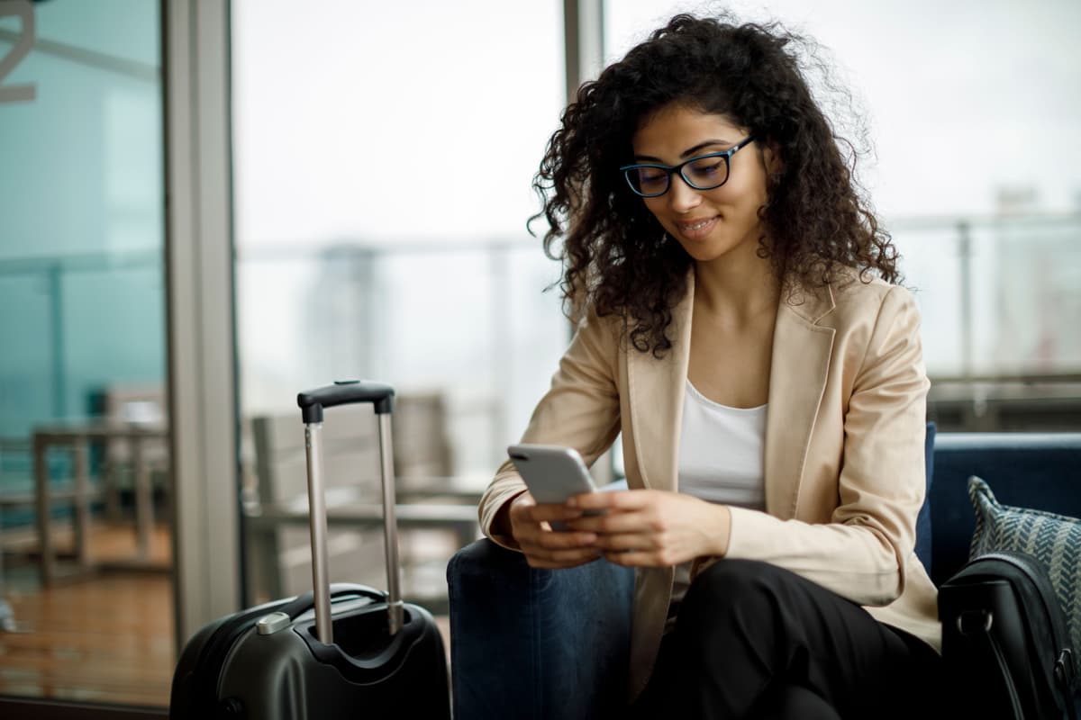 A woman with curly hair and glasses sits in an airport lounge, looking at her phone with a suitcase nearby.