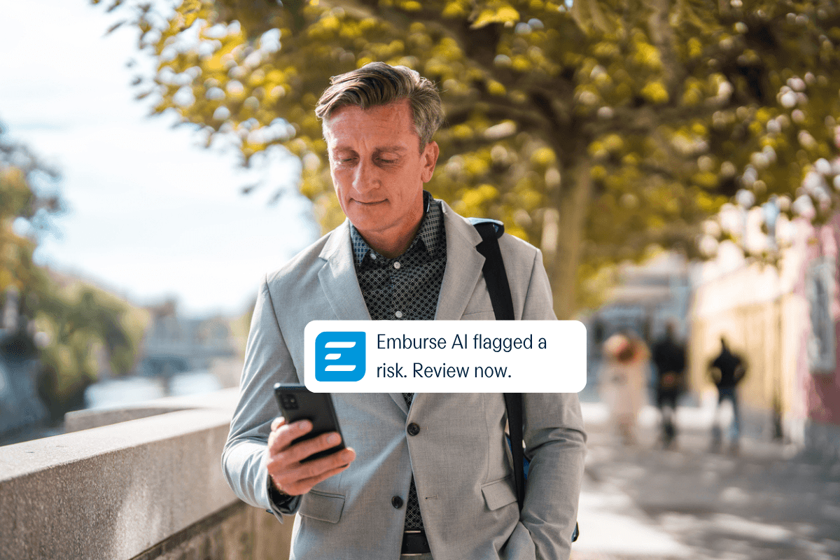 A man outdoors looks at his phone with a notification: "Emburse AI flagged a risk. Review now."