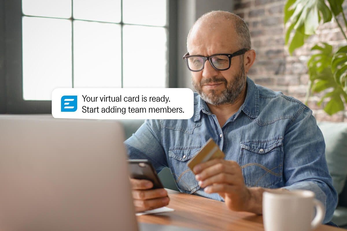 A man holds a credit card while looking at his phone, which displays a notification: "Your virtual card is ready. Start adding team members."