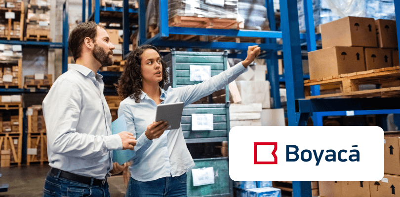 Two logistics workers in a warehouse, a woman pointing while holding a tablet, with the Boyacá logo.