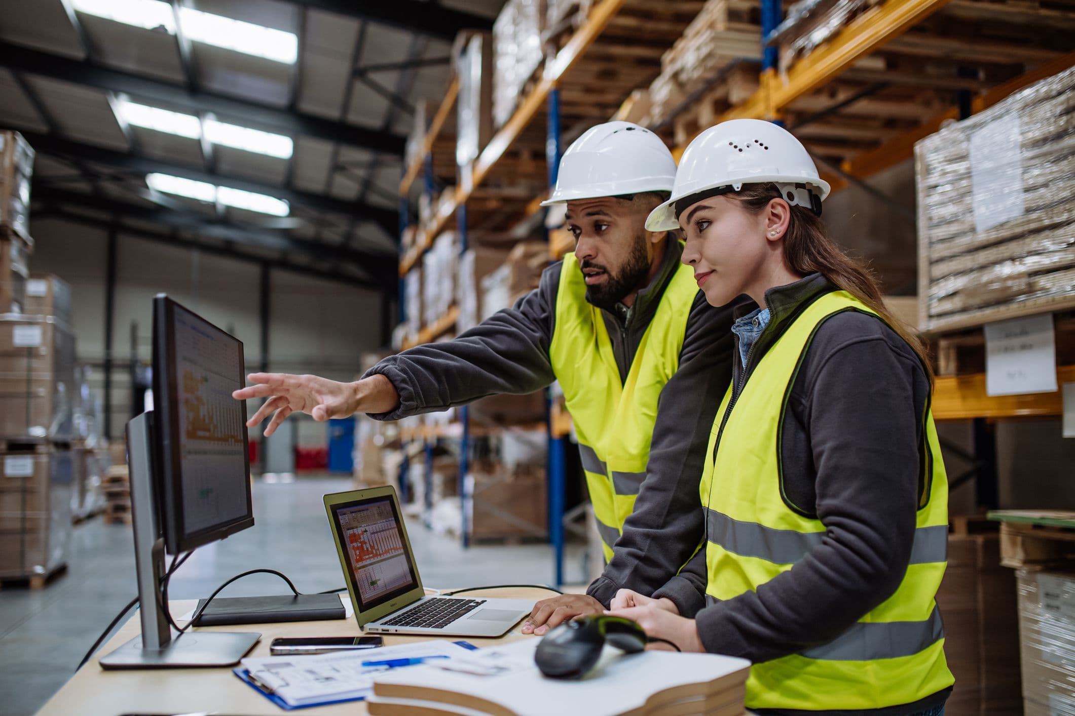 Two warehouse workers in hard hats and safety vests review data on a computer monitor and laptop.