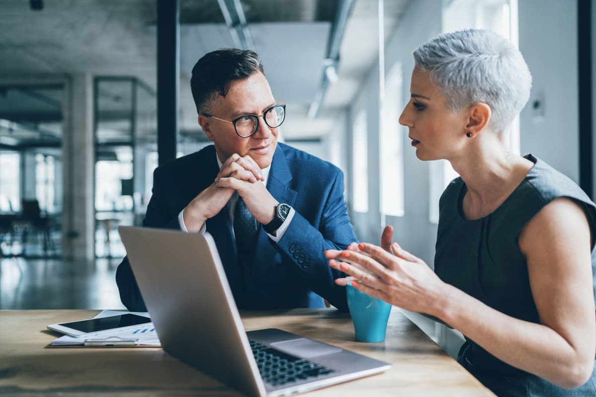 Two business professionals in a modern office, one speaking and one listening intently at a desk with a laptop.