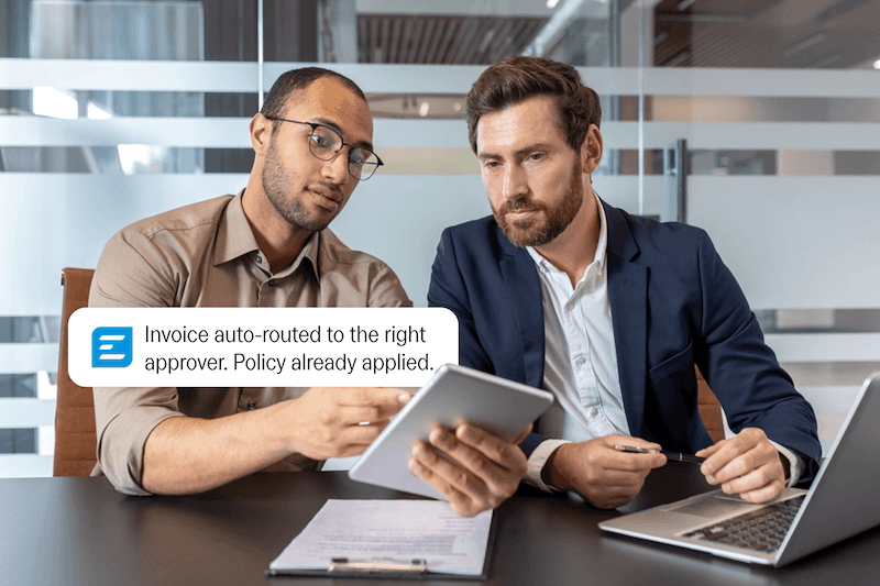 Two businessmen discussing information on a tablet, with a notification reading: "Invoice auto-routed to the right approver. Policy already applied."